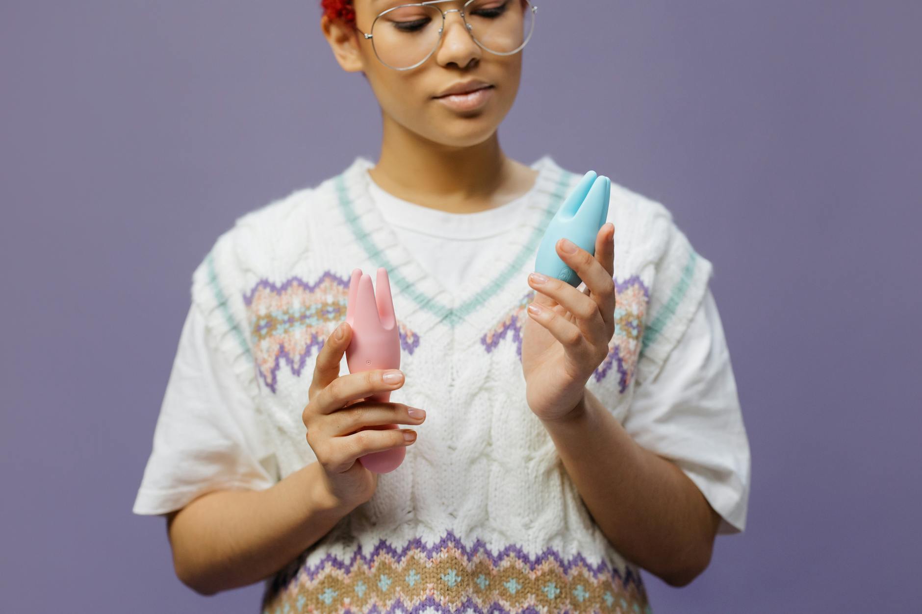 Woman with eyeglasses holding blue and pink silicone vibrators in a contemplative manner.